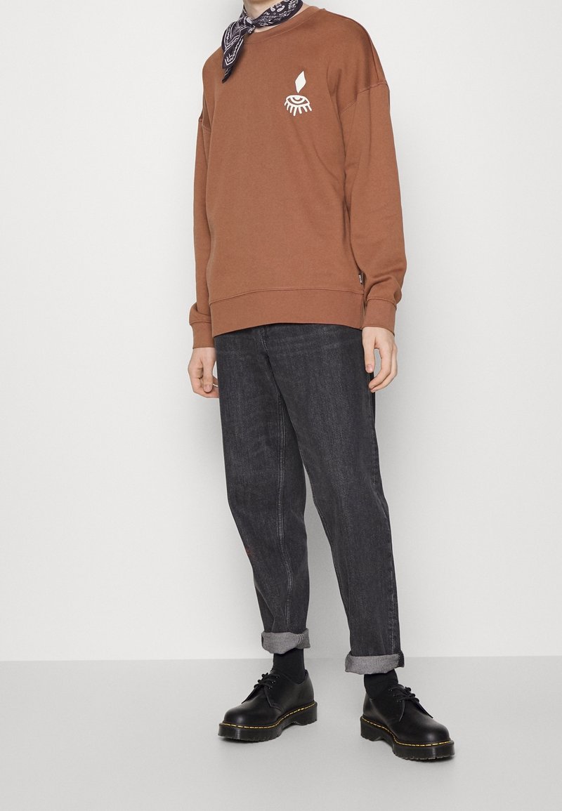 Personne portant un sweat-shirt marron avec un motif œil blanc, un jean noir retroussé, des chaussures noires et un foulard noir à motif autour du cou, debout contre un mur blanc.