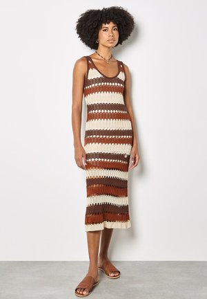 Mujer con cabello rizado que lleva un vestido de punto sin mangas hasta la rodilla con franjas horizontales en marrón, beige y crema, y sandalias planas marrones.