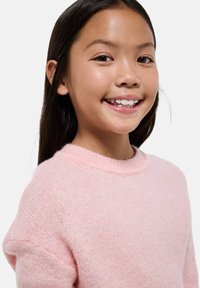 Jeune fille aux longs cheveux bruns, souriant, portant un pull en laine rose pâle sur fond clair.