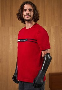 Punane puuvillane T-särk musta ja valge horisontaalse triibuga riba, millel on "TOMMY HILFIGER" tekst. Mustad kindad katavad proteeskäed.