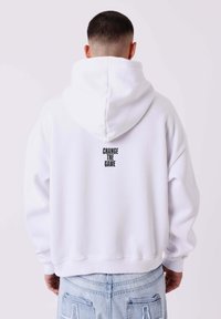 Sudadera blanca de corte holgado y con una capucha grande en la parte posterior. Presenta texto negro "CAMBIA EL JUEGO" en una disposición vertical. Material de algodón suave.