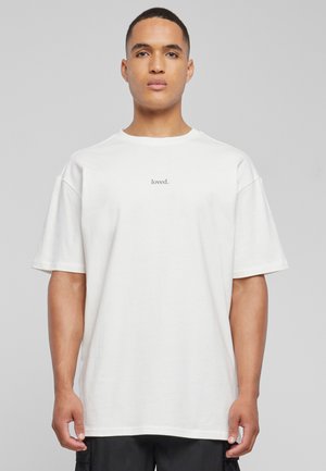 T-shirt oversize blanc en coton, à manches courtes, affichant le mot "loved." imprimé en petite police noire sur la poitrine.