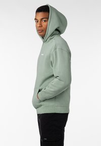 Hellgrüner Kapuzenpullover mit einer Kängurutasche und gerippten Bündchen. Logo auf der Brust, weiche Textur, lässige Passform. Trägt man mit schwarzen Hosen.