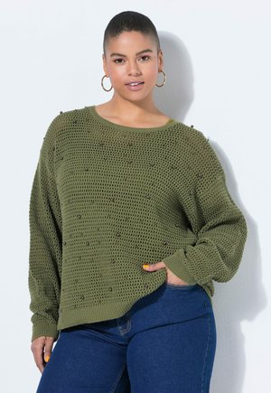 Femme aux cheveux courts portant des boucles d'oreilles créoles, un pull en maille vert olive avec des détails en perles, et un jean bleu, posant avec la main dans la poche.