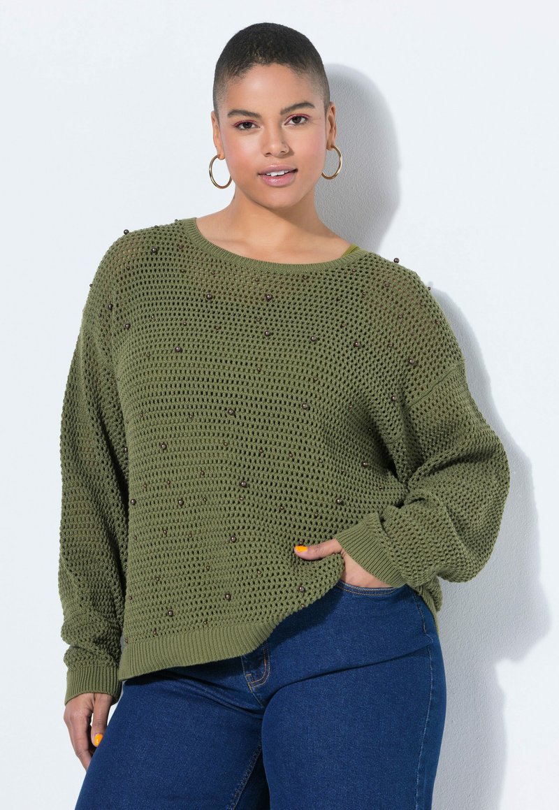 Donna con capelli corti che indossa orecchini a cerchio, un maglione verde oliva lavorato a maglia con dettagli di perline e jeans blu, posando con la mano in tasca.