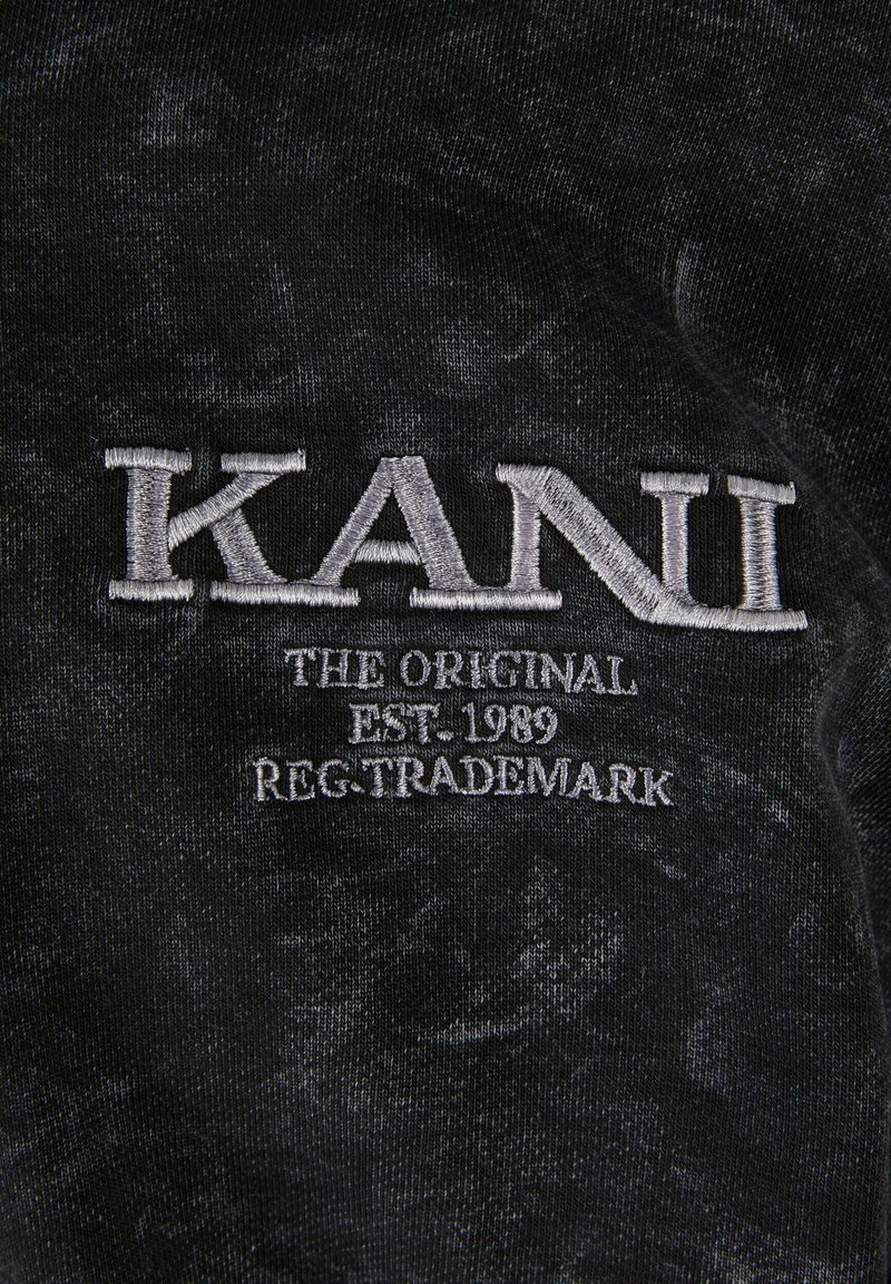 Tessuto nero caratterizzato da un aspetto testurizzato e sbiadito. Logo ricamato "KANI" in bianco con "THE ORIGINAL EST. 1989 REG. TRADEMARK" sotto di esso.