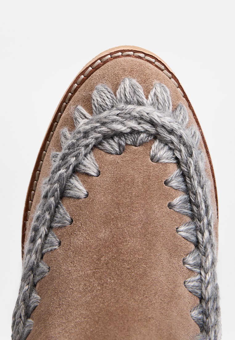 Zapato de mocasín de ante marrón con costuras trenzadas grises y bordes ondulados en la parte superior. Textura suave y punta redondeada.