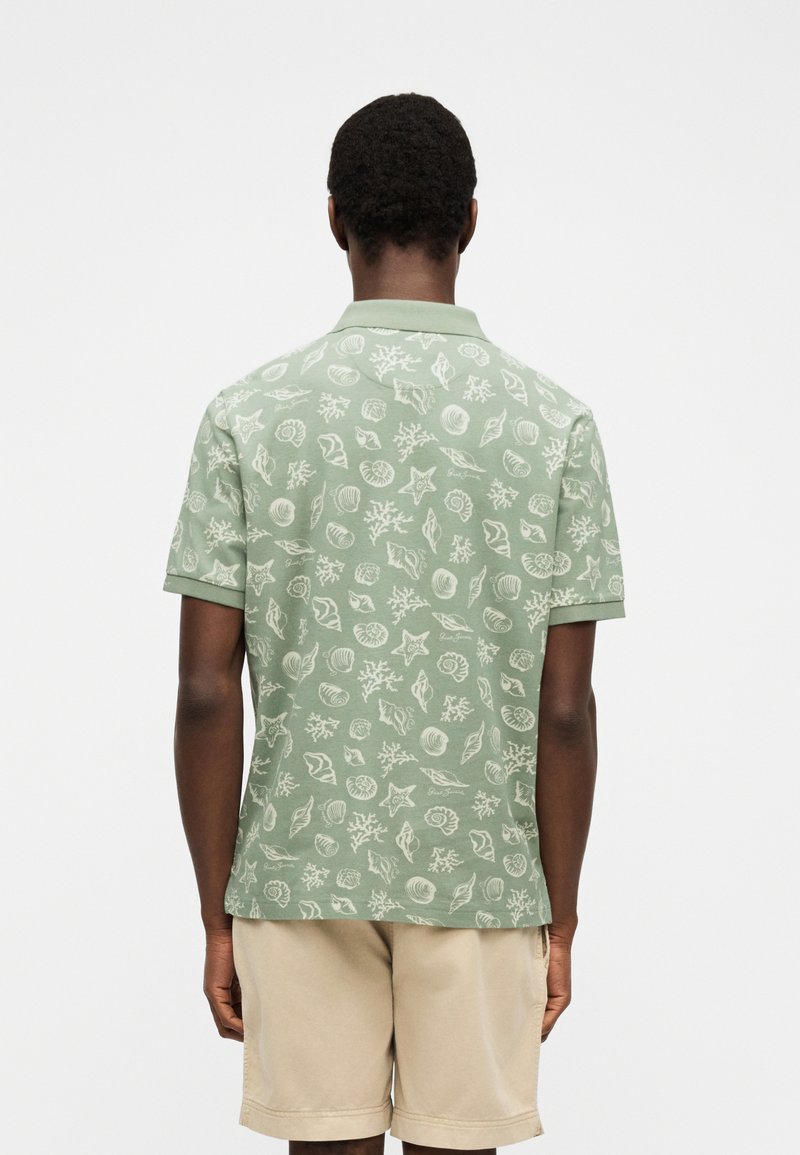 Uomo che indossa una polo a maniche corte verde chiaro con motivi di conchiglie e coralli bianchi, abbinata a pantaloncini beige, visto di spalle su sfondo bianco.