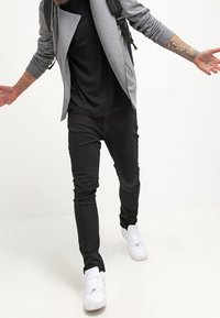 Chaqueta gris con cremallera frontal sobre una camisa negra, combinada con pantalones negros y zapatillas blancas. La chaqueta presenta un diseño asimétrico único.