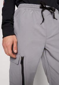 Světle šedé sportovní kalhoty s elastickým pasem, přední kapsou na zip, boční kapsou a šňůrkou na stažení. Hladká textura, funkční design.