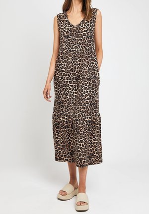 Vrouw draagt een mouwloze, V-hals, kuitlange jurk met luipaardprint en een geplooide taille, gecombineerd met beige platform slippers, staand tegen een witte achtergrond.