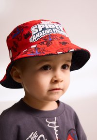 Cappello a secchiello rosso con grafiche e loghi di Spider-Man, con fodera interna nera. La maglia grigia del bambino presenta una scritta bianca.