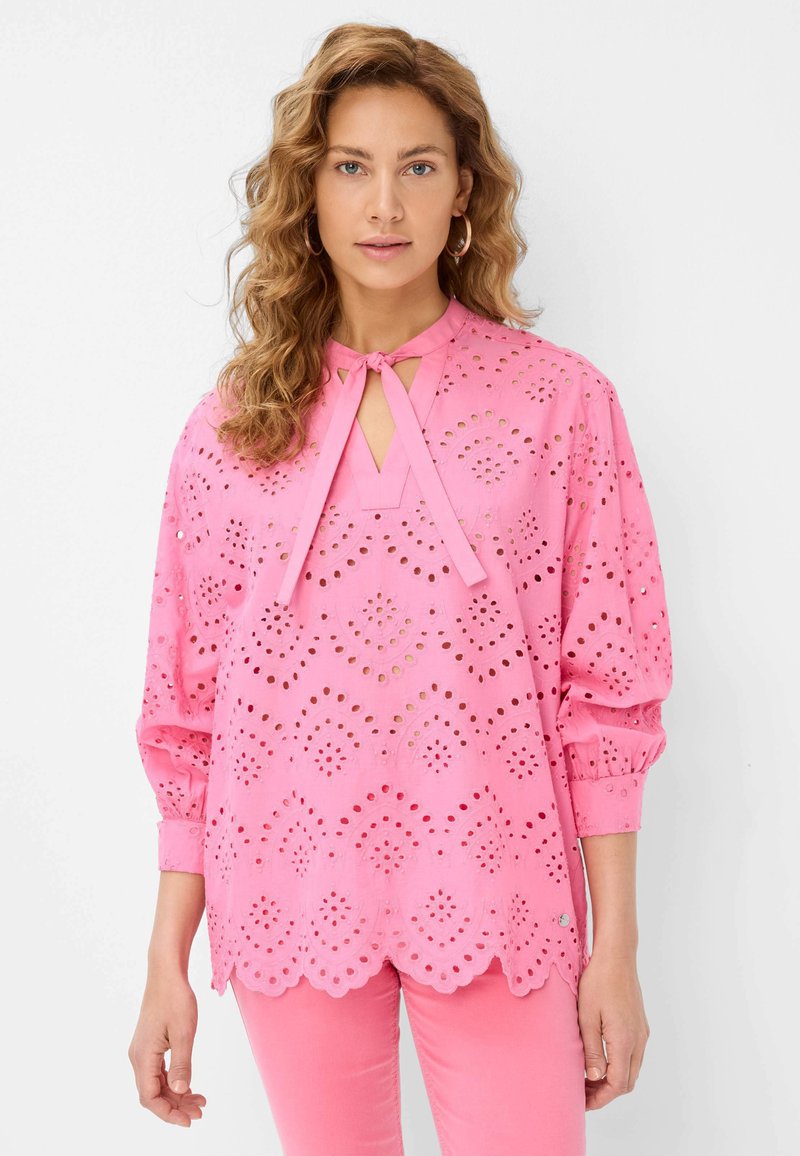 Blusa rosa con motivo floreale a intaglio, chiusura a fiocco e maniche lunghe. Presenta un orlo smerlato e dettagli ricamati.