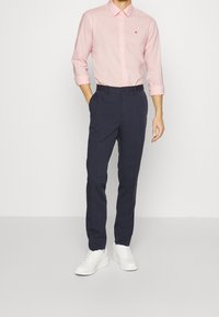 Camicia button-down rosa chiaro con maniche arrotolate, pantaloni navy e sneakers bianche. La camicia presenta un piccolo logo rosso sul petto.