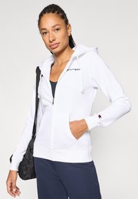 Λευκό φούτερ με φερμουάρ, κατασκευασμένο από μαλακό ύφασμα, με τσέπη μπροστά, κεντημένο λογότυπο και κουκούλα με κορδόνι. Φορεμένο με σκούρο παντελόνι.