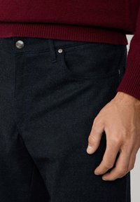 Jean en denim bleu foncé avec une poche avant, des coutures contrastantes et un bouton en argent. Le tissu présente un aspect texturé.