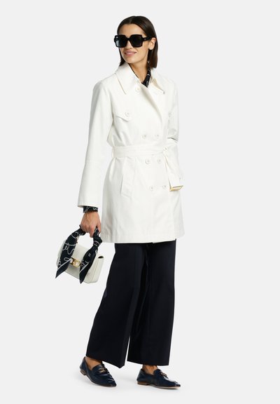 Manteau trench blanc à double boutonnage avec ceinture, associé à un pantalon large bleu marine et des mocassins noirs. Les accessoires incluent des lunettes de soleil noires et un sac à motifs.