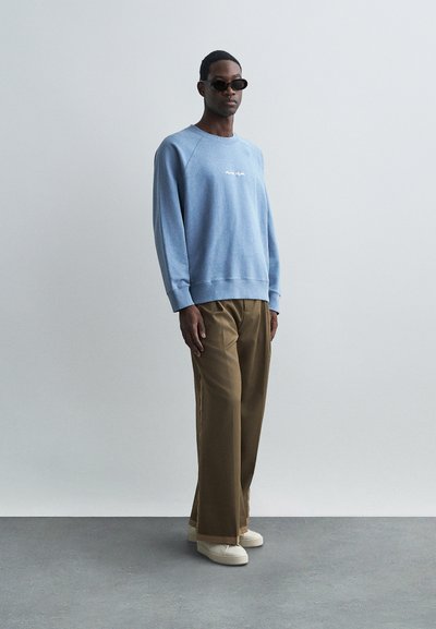 Sudadera azul claro con cuello redondo, logo bordado y puños acanalados. Combinada con pantalones sueltos color beige y zapatillas blancas. Gafas de sol negras.