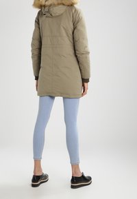 Osoba stojící zády, oblečená v zelené parce s kožíškem na kapuci, světlých modrých úzkých džínách a černých platformových botách na jednobarevném pozadí.