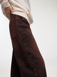 Pantaloni in denim marrone a gamba larga con molte tasche e una finitura testurizzata, abbinati a una felpa crema con tasca anteriore.