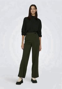 Femme debout portant un chemisier noir à manches longues, un pantalon large taille haute vert olive, des chaussettes beiges et des mocassins noirs.