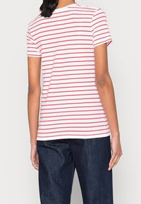 Person iført en hvid kortærmet skjorte med røde vandrette striber og mørkeblå jeans, vist fra ryggen.