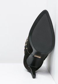 Zapato de charol negro de tacón alto con punta afilada, con tachuelas de color dorado, suela texturizada y diseño de tacón stiletto.