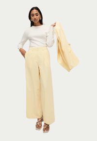 Lichtgele wijde broek met plooien en zakken aan de zijkant, gedragen met een aansluitend wit ribgebreid long-sleeve shirt. Een bijpassende jas wordt vastgehouden.