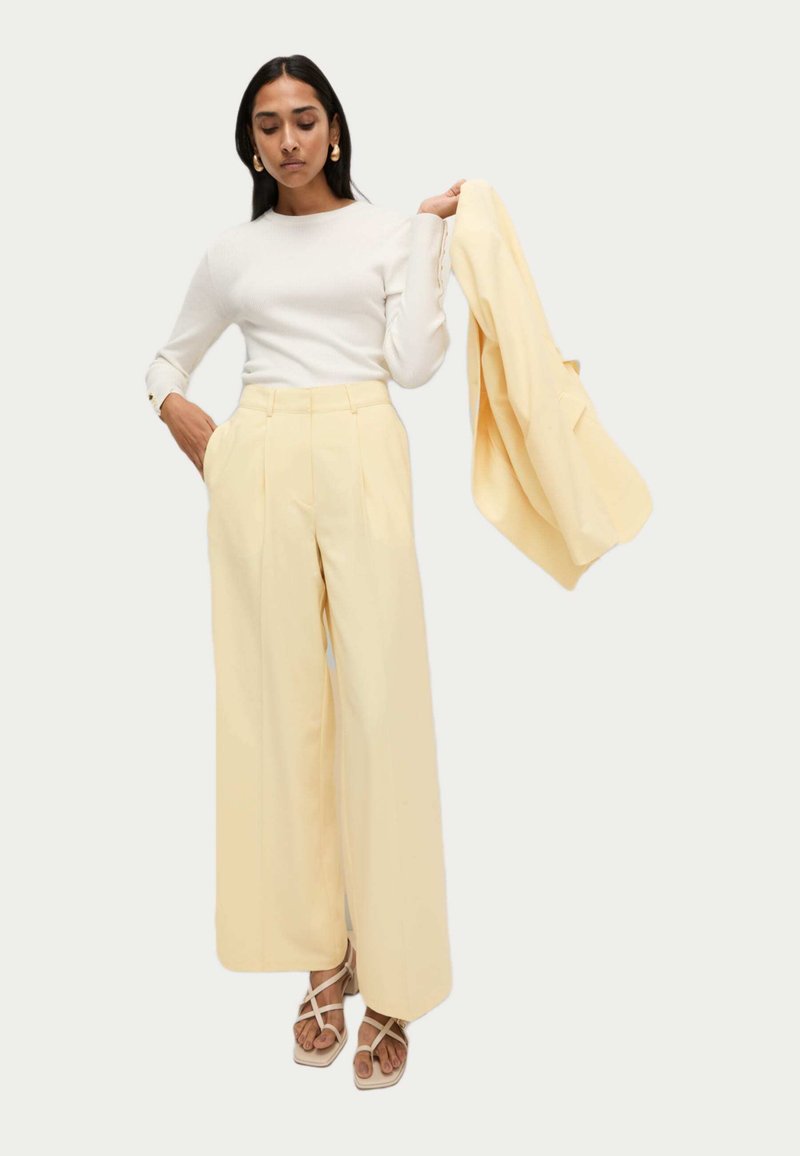 Lichtgele wijde broek met plooien en zakken aan de zijkant, gedragen met een aansluitend wit ribgebreid long-sleeve shirt. Een bijpassende jas wordt vastgehouden.