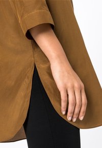 Brun, semi-strukturerad blus med uppkavlade ärmar, en böjd nederkant och en sidoflik, kombinerad med svarta, åtsittande byxor.