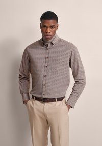 Button-up shirt featuring a brown and cream geometric pattern, long sleeves, and brown buttons, paired with light beige pants.