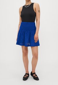 Jupe bleu dentelle avec un ourlet à volants, un motif floral texturé et une taille élastique. Associée à un haut noir sans manches et des chaussures noires.