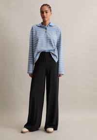 Chemise rayée bleue et blanche à manches longues avec un col, associée à un pantalon large noir et des chaussures blanches avec une semelle marron.