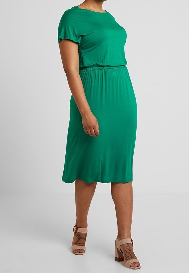 Femme debout dans une robe verte longueur genou à manches courtes, portant des sandales beiges à talon bloc ouvertes à l’avant, avec des brides à la cheville.