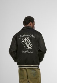 Vīrietis melnā jakā ar uzrakstu "Flatbush Ave. KANI Est 1989 Bk. New York" aizmugurē, valkājot zaļas bikses ar kabatas logotipu, stāv uz vienkārša fona.