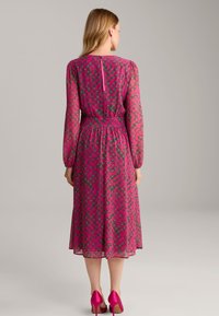 Langärmliges pinkes Kleid mit einem wirbelnden Muster in dunkleren Farben, tailliertem Bund und einem Reißverschluss hinten. Getragen mit fuchsienfarbenen High Heels.