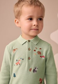 Polo a maglia verde chiaro con bottoni marroni e ricamo colorato di animali, tra cui una volpe, un riccio e dei funghi.