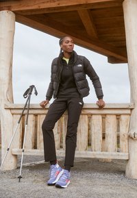 Giacca a vento nera, maglia nera a maniche lunghe, pantaloni neri e sneakers blu con accenti rossi; bastoni da trekking accanto a una struttura di legno.