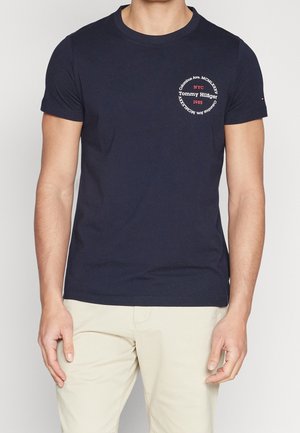 Man wearing navy blue short-sleeve T-shirt with circular white and red Tommy Hilfiger logo on left chest and beige pants.