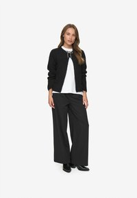 Black knitted cardigan with long sleeves, white top underneath, and wide black pants, worn with black pointed-toe shoes. Simple design.