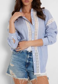 Camicia a righe blu e bianche con bordo in pizzo, presenta maniche arrotolabili e una vestibilità comoda, abbinata a pantaloni shorts in denim consumati.