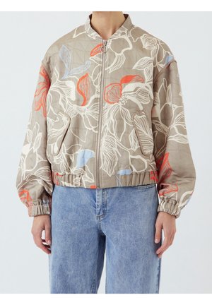 Personne portant une veste beige zippée avec de grands motifs floraux blancs, rouges et bleus et des poignets élastiques, associée à un jean bleu clair.