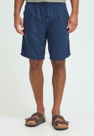 Marineblauwe short met trekkoord in een ontspannen pasvorm, voorzien van zijzakken en een gladde, lichte stof. Draagt met bruine sandalen.