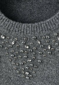 Suéter gris de punto con un cuello acanalado y pequeños embellishments metálicos dispersos de varias formas y tamaños en la parte frontal.
