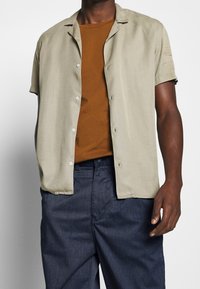 Ljusbeige kortärmad skjorta med knappstängning, lagd över en brun t-shirt, i kombination med marinblå byxor. Slät tygstruktur.