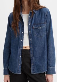 Donna che indossa una camicia di jeans blu con bottoni sopra un top corto bianco e pantaloni neri, con lunghi capelli castani in vista.