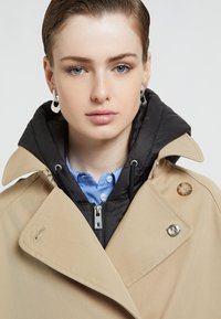 Beige trench coat dengan tudung berlapis fleece hitam, dilengkapi dengan zip perak, butang bulat, dan baju berjalur biru berkolar di bawahnya.