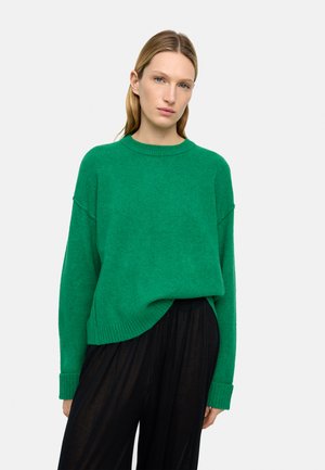 Kobieta o długich blond włosach, ubrana w jasny zielony sweter z dzianiny i luźne czarne spodnie, stojąca na tle jednolitego jasnego tła.