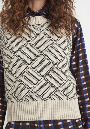 Femme portant un gilet en tricot crème avec des motifs en zigzag et chevrons noirs par-dessus une chemise à carreaux bleue, marron et blanche à manches longues.