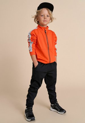 Kind steht mit in den Taschen steckenden Händen da, trägt eine orangefarbene Jacke mit Reißverschluss, schwarze Hose, schwarze Turnschuhe und eine schwarze Mütze.